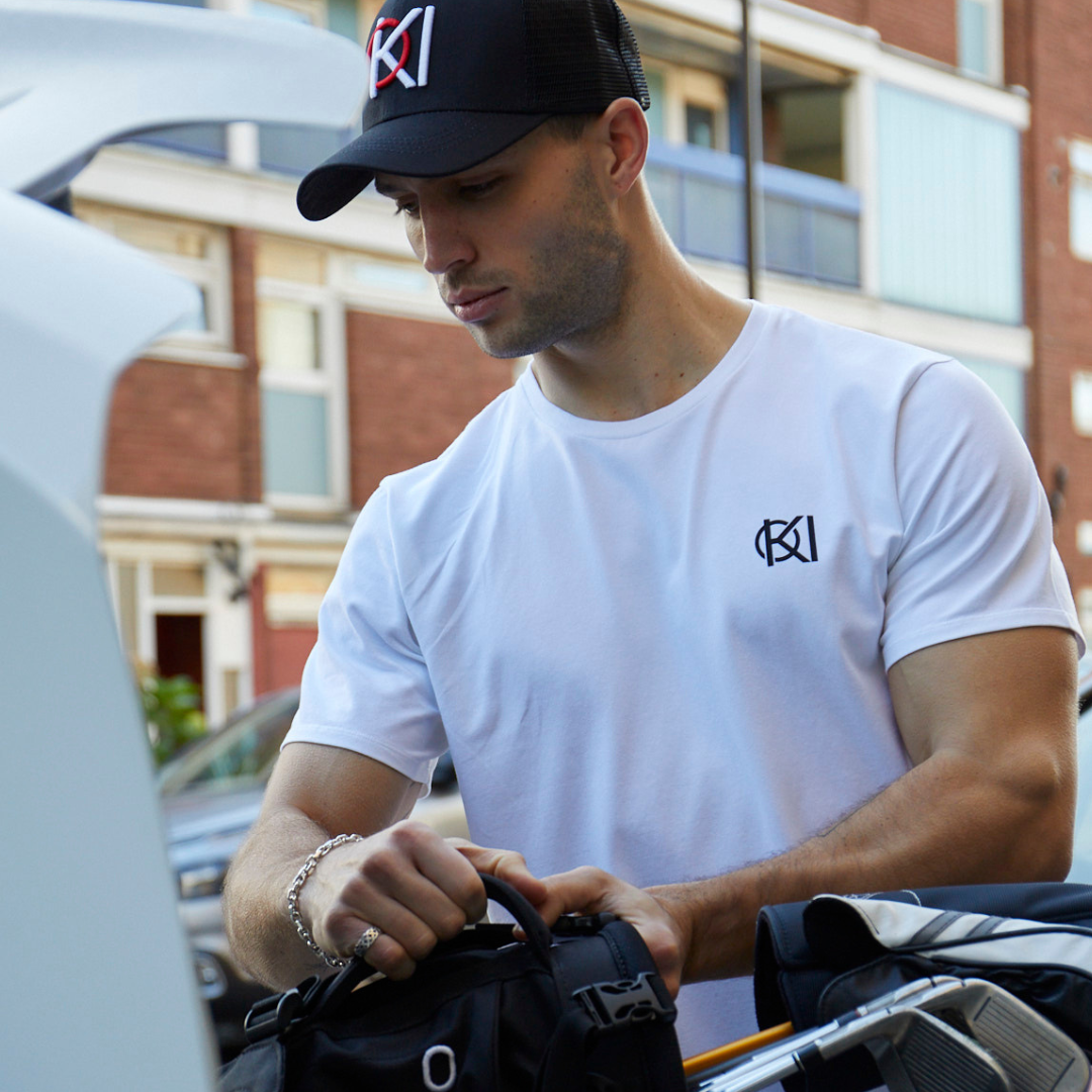Man wearing a white t-shirt and black cap with a logo, standing outdoors.