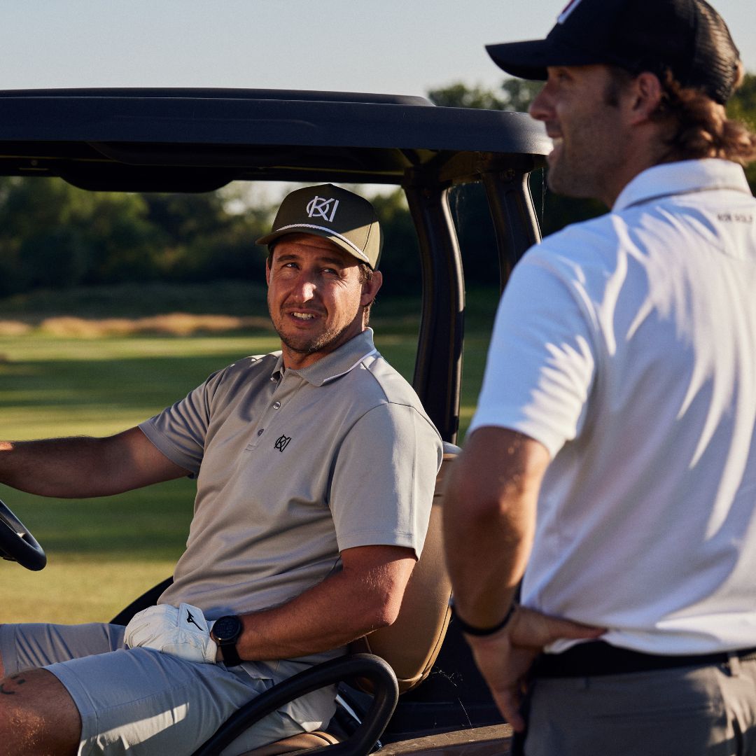 Two men in golf attire sitting in a golf cart on a golf course.