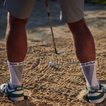 Person in golf shoes and socks standing on a sand trap with a golf club and ball.