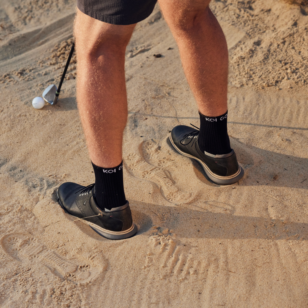 Person standing in a bunker wearing black golf socks with visible branding