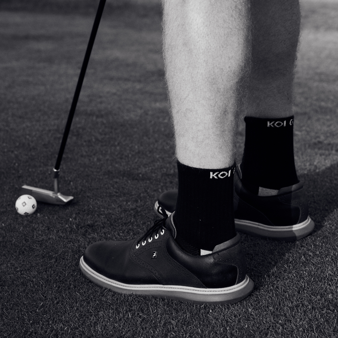 Person on a golf course wearing black shoes and golf socks with 'Koi' branding.